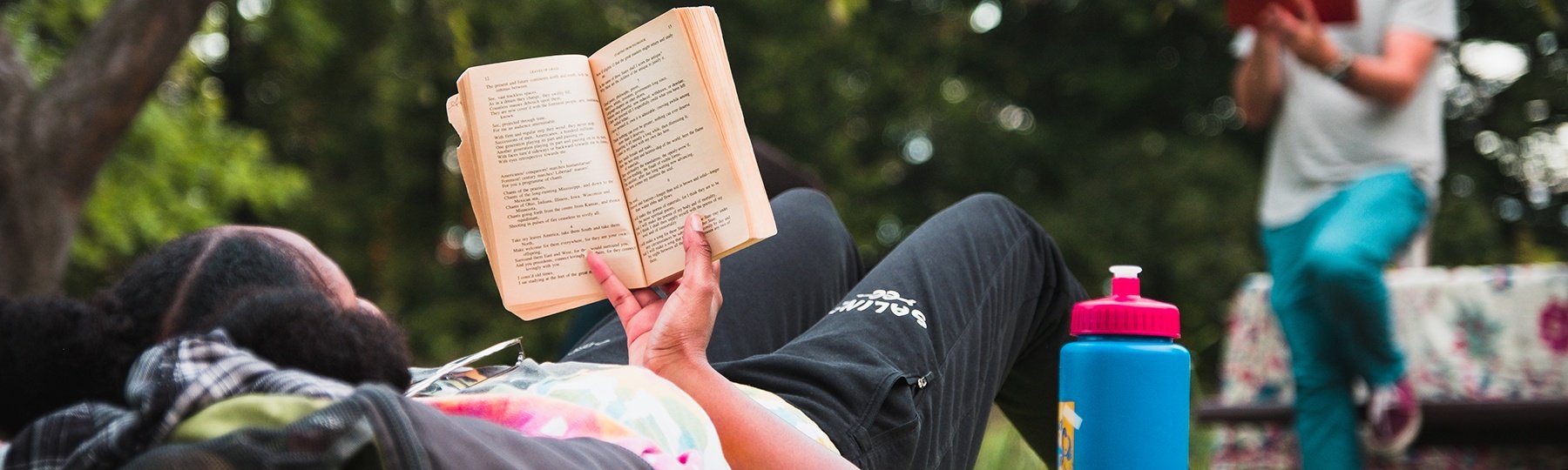 Student seated on the grass holds a book in her hands and reads while other students sit nearby to listen.