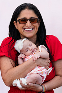 Dr. Mona Hanna wearing a red shirt and tinted glasses holds a baby wearing a white headband and white floral outfit.