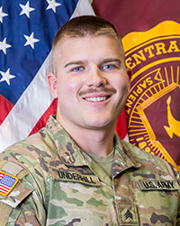 Closeup image of SGT Trevor Underhill wearing his military uniform with the American and Central Michigan University flags behind him.