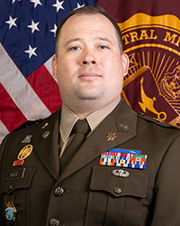 Closeup image of MAJ Christopher Mace wearing his military uniform with the American and Central Michigan University flags behind him.