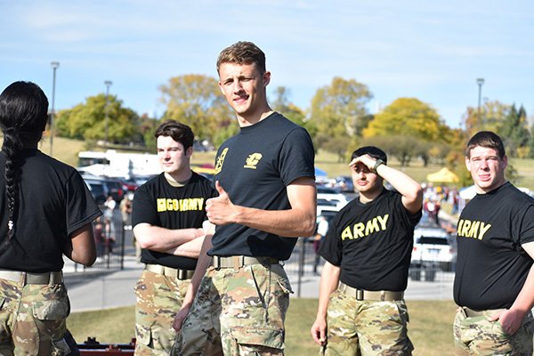 An Army ROTC student wearing a black t-shirt and green camouflage pants gives a thumbs up while other students stand behind him.