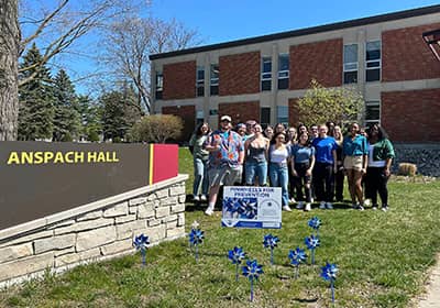A group of college students standing behind a child prevention sign and pinwheels in front of CMU's Anspach Hall.