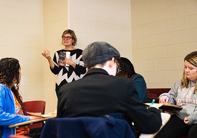 JoEllen DeLucia stands in a classroom holding a book with students seated in desks around her