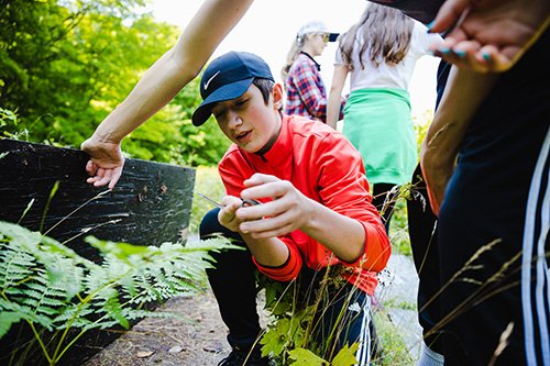 A high school student in a red jacket and blue baseball cap holding a small snake.