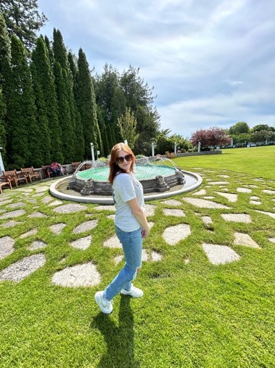 A woman in jeans and a t-shirt standing in front of a fountain surrounded by grass and trees.