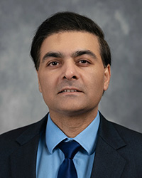A man in a dark suit and blue shirt in front of a grey background.