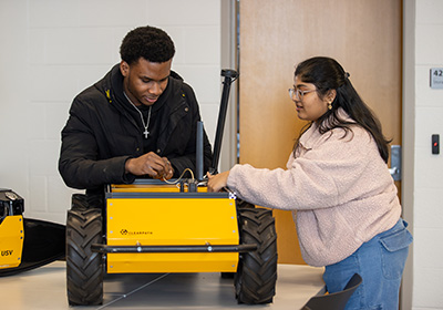 Two students collaborate while working on a yellow four-wheeled robotic vehicle in a classroom lab setting.