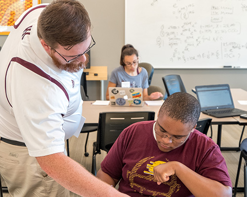 A man looking pointing to a math equation in aman in a red shirt's notebook.