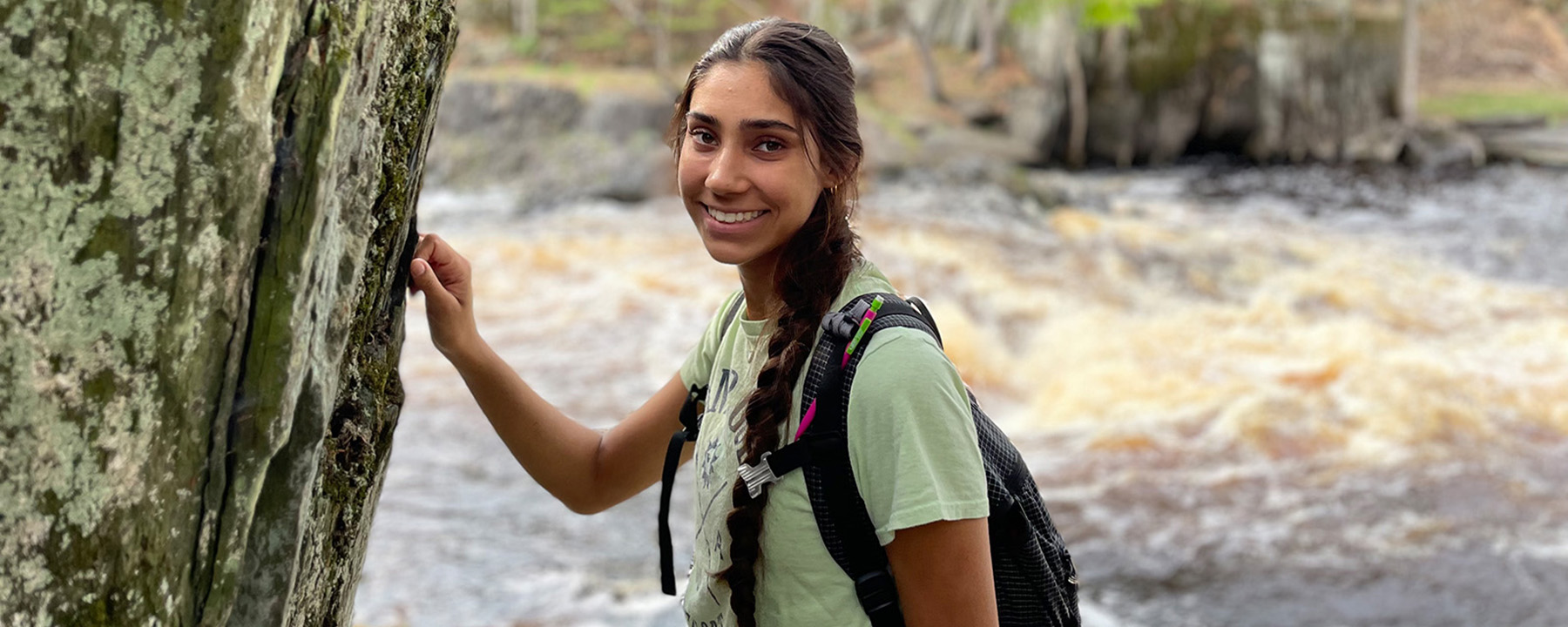A woman with a backpack standing in front of a river with her hand against a rock cliff.