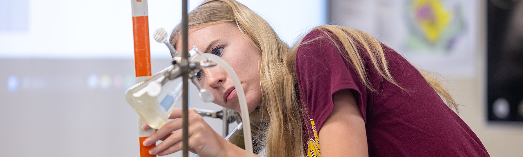 A woman in a maroon t-shirt examining a sample in a vial suspended from a vertical rod.