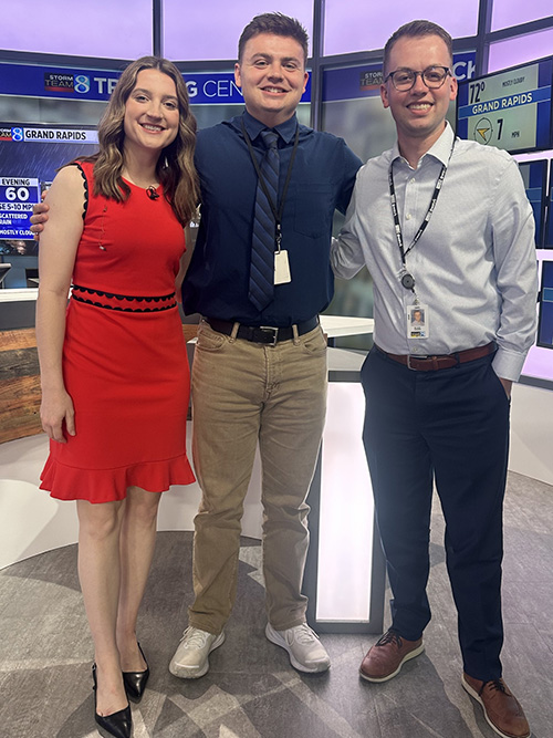 A student intern stands smiling with two WOOD TV8 team members inside the station’s weather center.