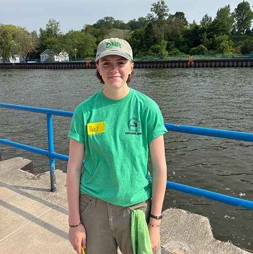 A student in a green t-shirt and brown hat standing on a dock in front of a large body of water.