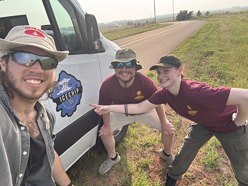 Three students smile in front of a white van marked with the ICECHIP field campaign logo.