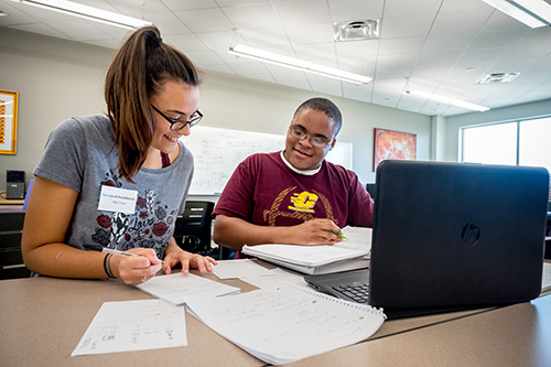 Two students at a table with a bunch of papers and a laptop on it.