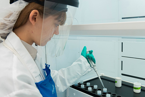 A woman in a lab coat and protective face shield preparing samples in a lab environment.