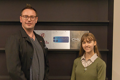 A man with glasses and a student with long hair and a green sweater stand in front of a sign that says STAR LAB.