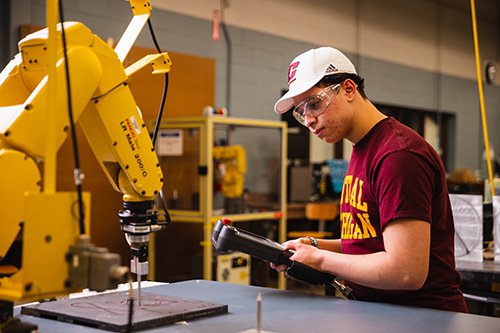 A CMU student wearing safety glasses and a Central Michigan University shirt and cap operates a robotic arm in a lab, using a handheld controller to program its movements.