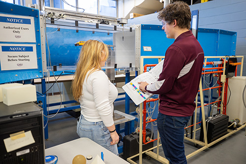 Two students looking at procedures in a manual before running a test in a small wind tunnel enclosure.
