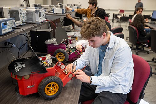A student in a light blue jacket works on a robotic vehicle in an engineering lab, adjusting its wiring and components. 