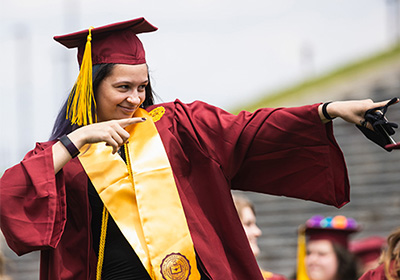 A student dress in a maroon cap and gown with a gold stole points both index fingers to the left and smiles.