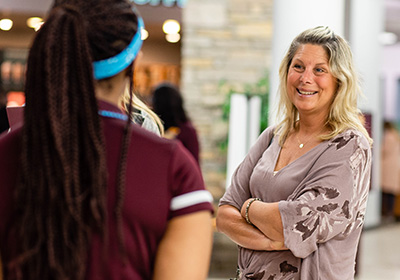 A student in a maroon polo stands with their back to the camera talking to a parent. The parent, wearing a light purple blouse, stands with her arms folded looking at the student and smiling.