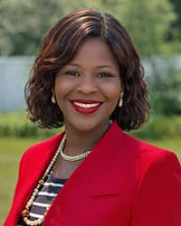 Kechinyere Iheduru-Anderson, wearing a red blazer, black and white striped blouse, and gold necklaces, stands outside in a garden and smiles at the camera.