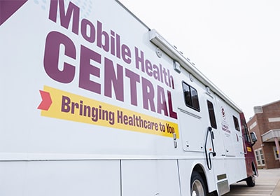 The white Mobile Health Central bus with Mobile Health Central painted in maroon and Bringing Healthcare to You in maroon on a yellow banner. The bus is parked in the parking lot next to the College of Health Professions.