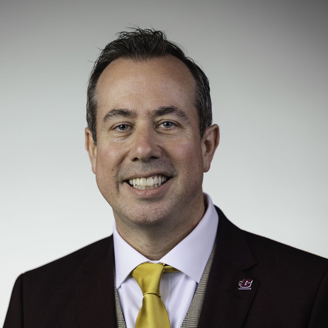 President-Elect Neil MacKinnon wears a maroon suit with a gold tie while posing for a professional head shot against a grey background.