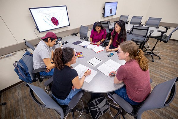 A group of students work together at an active classroom table. They are wearing maroon shirts and have open notebooks in front of them.