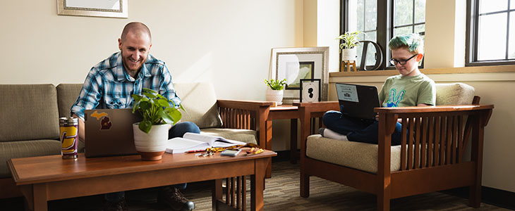 Father taking online class on his computer while young son sits nearby doing his homework on his computer.
