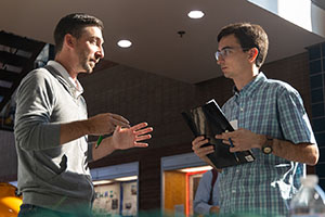 A man wearing a gray sweater emphasizes with his hands while talking to a younger man in a checked button-down shirt and glasses.