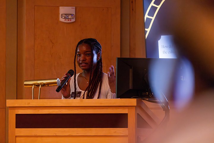A female student holds a microphone while standing at a wooden podium that has a gold reading light attached to it.