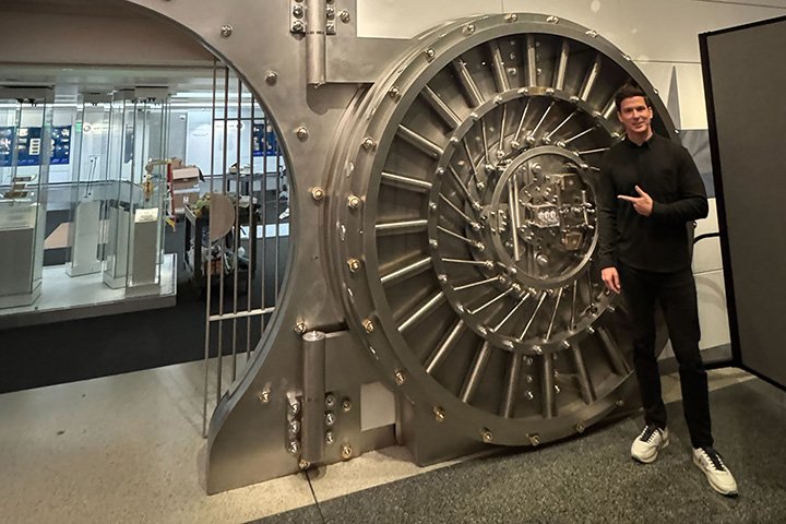 A man in sneakers and a dark track suit stands next to and points to an open bank vault leading to a museum exhibit.