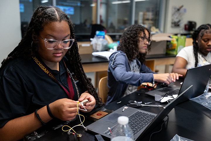 A young woman wearing a dark shirt and glasses examines an electronic circuit.