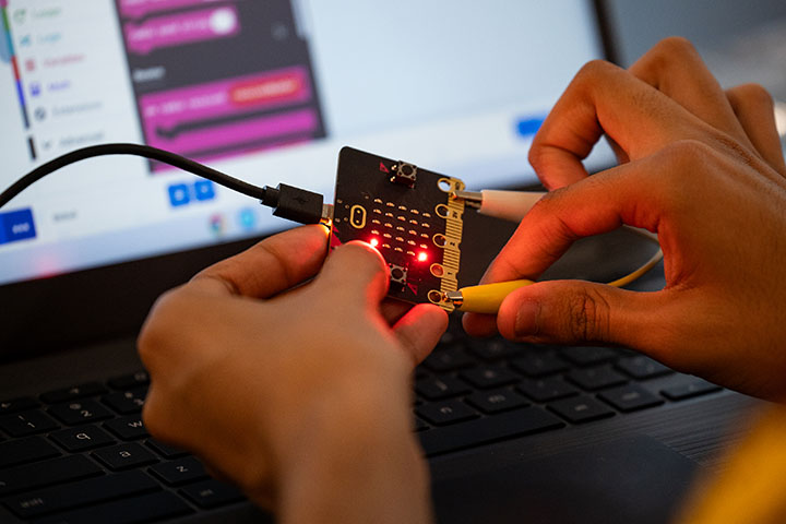 One hand holds a digital circuit board while another hand touches it with a test equipment lead.