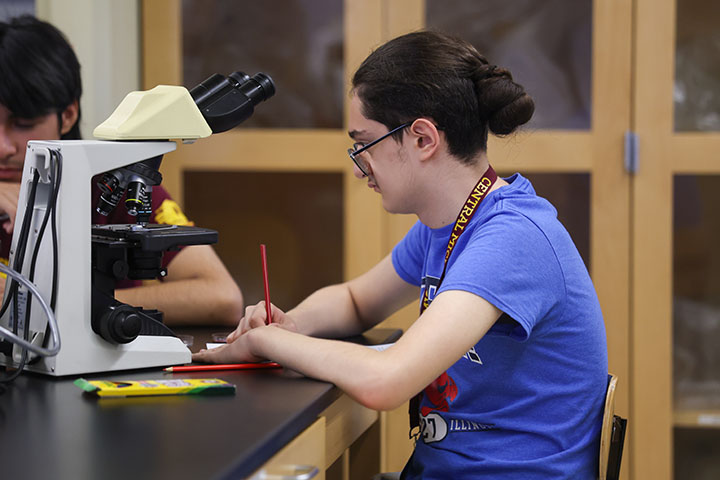 A young man wearing glasses and a blue T-shirt writes notes on a piece of paper with a red pen.