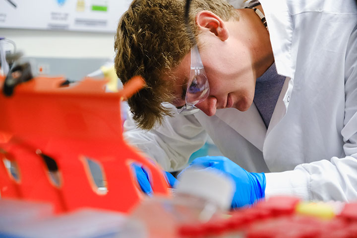 A man in safety glasses, a white lab coat and blue gloves concentrates on creating samples for analysis in a lab.
