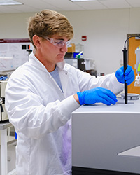 A young man in safety glasses, a white lab coat and blue gloves inserts a sample into a piece of lab analysis machinery.