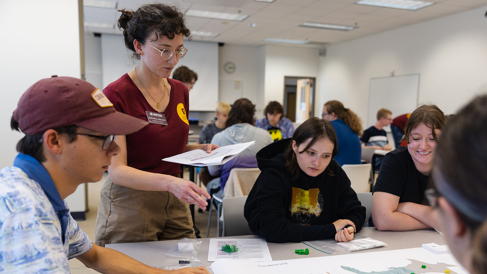 A woman wearing glasses and with black curly hair pulled back into a bun holds a packet of papers in one hand and points at a gaming map with the other while three people sitting at a table look on.
