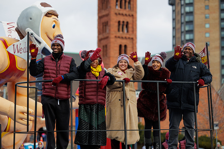 Five people stand on top of the CMU parade float waving to the crowd