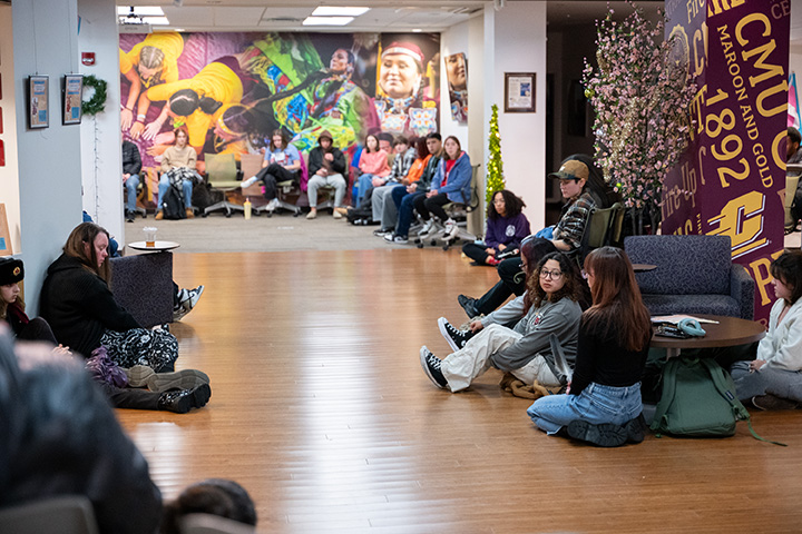 A large group of students sit along the ground and in chairs inside a room
