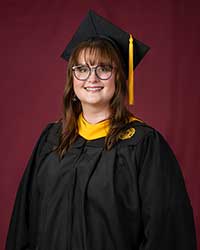 A dark haired woman in a graduation cap and gown stands in front of a maroon backdrop
