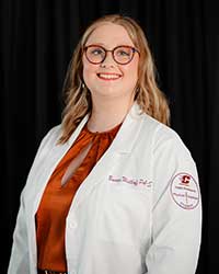 A blond woman wearing glasses and a physician assistant white coat stands in front of a dark background