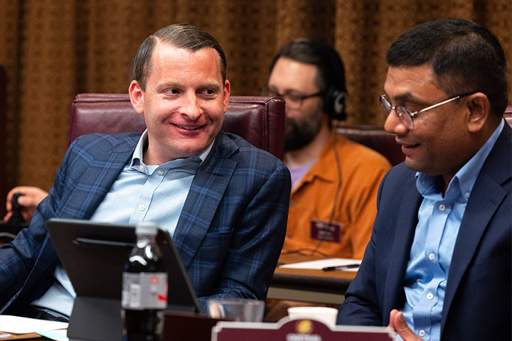 Two men wearing suit jackets and blue shirts sitting at a table having a discussion.