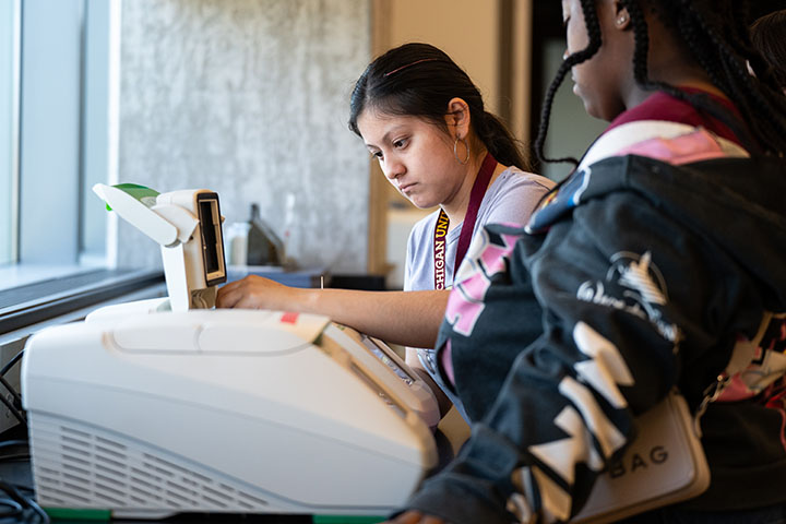 A young woman with a long ponytail puts a sample into lab equipment.