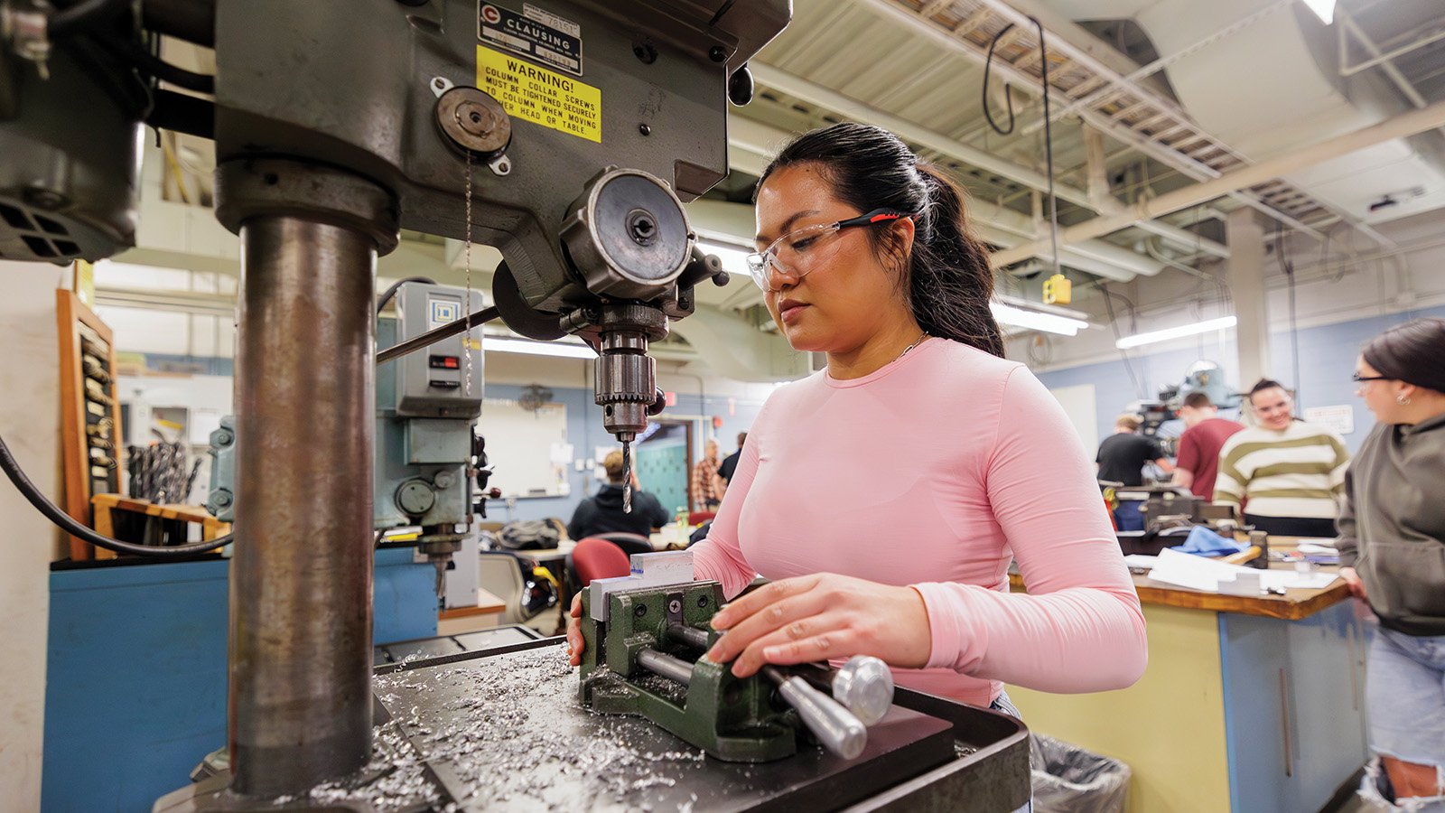 World Class_Engineering Female student wearing protective glasses stands at a machine inside an engineering lab.
