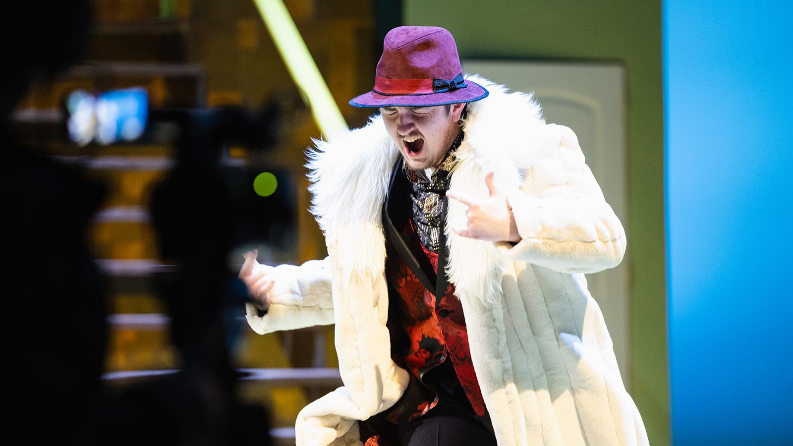 World Class_Performances A male student performing on stage wearing a faux fur lined coat and a bright purple hat.