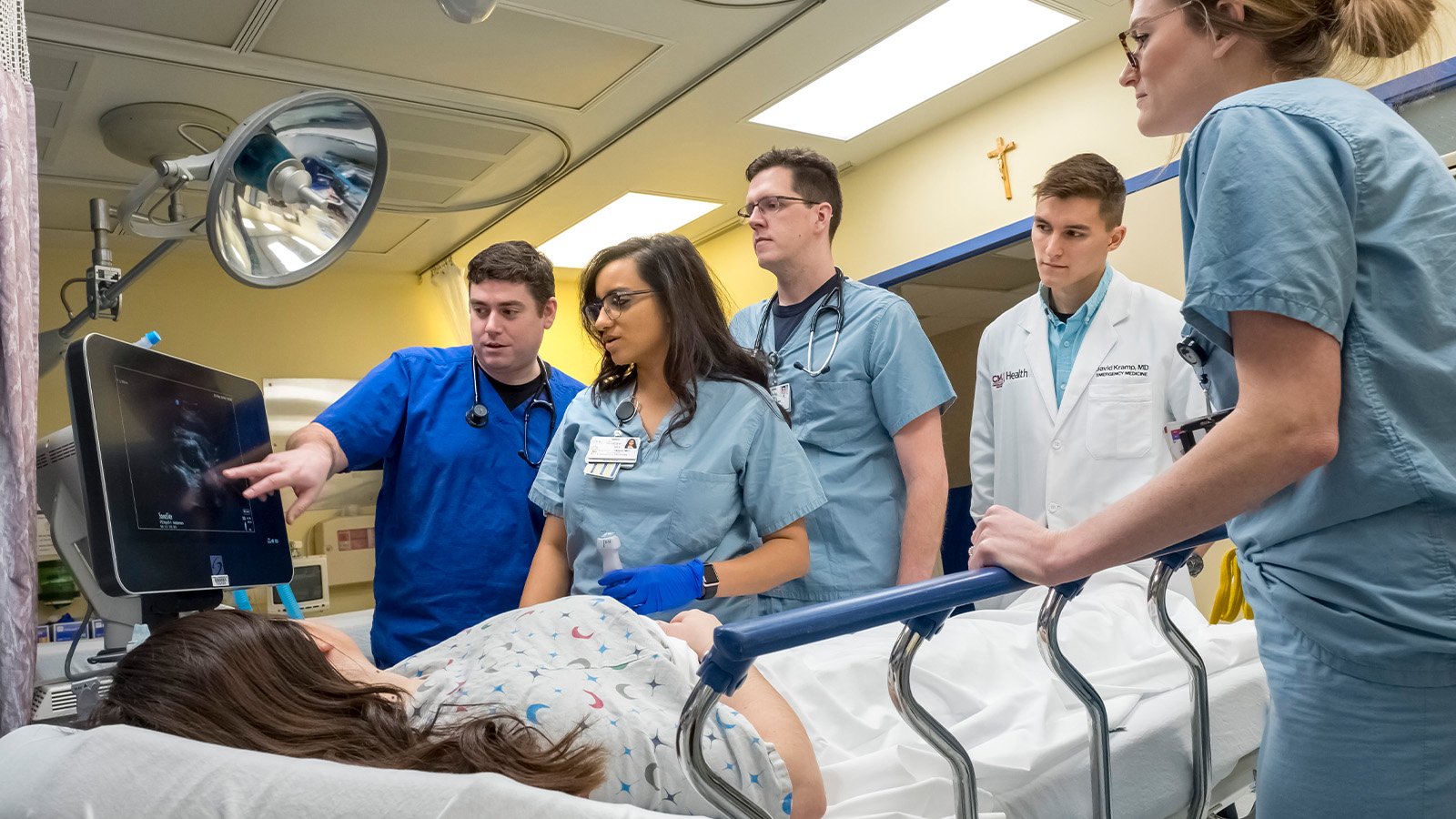 World Class_Residents Medical students stand around a patient in a hospital bed while they review data on a computer monitor.