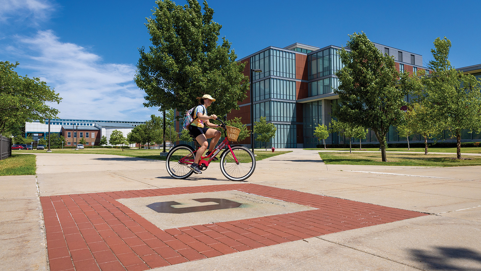 World Class_Sustainability A female rides a red bike across campus in the summer, passing in front of a large bioscience building.