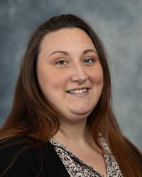 Brittany Yenior, Administrative Secretary,  profile portrait. She has long brwon hair, wearing a black cardigan over a white and black floral top and a grey background for the picture